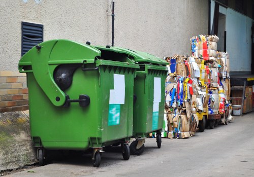 A row of two large, bright green wheelie bins with closed lids positioned on a concrete pavement beside a beige building. The bins have white labels on their front and are placed close to a wall with a black ventilation grille partially visible on the left. To the right of the bins, there are multiple tightly packed bundles of flattened cardboard boxes and paper waste, some with visible blue and red markings. The cardboard is stacked against the building's exterior wall, which features a glass window and a partial view of a nearby door in an urban environment. The scene appears to be part of a rubbish removal service in Limehouse, with the waste materials prepared for collection or disposal. The pavement is clean, and the lighting suggests a daylight setting, creating a clear view of the waste materials and bin arrangement, aligned with professional rubbish clearance activities.