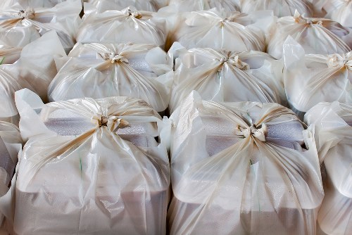 The image displays multiple beige or off-white plastic rubbish bags tightly knotted at the top, arranged closely together, filling the foreground of the photo. These bags appear to contain garden waste or general rubbish, with some showing slightly crumpled or shiny surfaces, indicating they are made of polyethylene. Behind the collection of bags, a grey vehicle or container may be partially visible, suggesting a rubbish collection or removal process. The environment seems to be outdoors, possibly on a driveway or pavement area, with natural daylight illuminating the scene, highlighting the texture of the plastic and the knots securing each bag. The setting aligns with waste management activities typical of Garden Clearance Limehouse, demonstrating the disposal or collection of garden refuse or household rubbish in an organized manner for clearance or recycling, in the local area of Limehouse, East London.