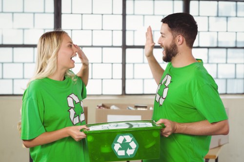 A photograph of two waste management workers indoors, both wearing bright green T-shirts featuring a recycling symbol on the front. The woman, with long blonde hair, is smiling and appears to be engaging in conversation with the man, who has dark hair and a beard. The man is holding a clear, green recycling bin with a white recycling logo on the side, and they are giving each other a high-five. In the background, there are large windows with a grid pattern, allowing natural light to illuminate the scene. The setting suggests an environment related to waste collection or recycling services, aligning with the activities of Garden Clearance Limehouse, a rubbish removal service specializing in clearance solutions within the local area near Limehouse and the London postcode E14. The overall scene conveys teamwork, environmental awareness, and professional waste management practices in a clean, organized indoor space.
