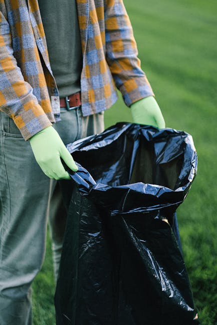 A person wearing a green gardening glove on each hand and dressed in a casual, plaid long-sleeve shirt over a green t-shirt, is holding open a large black rubbish sack made of plastic, which is lined 