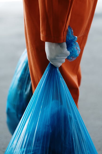 A person wearing a green gardening glove on each hand and dressed in a casual, plaid long-sleeve shirt over a green t-shirt, is holding open a large black rubbish sack made of plastic, which is lined 